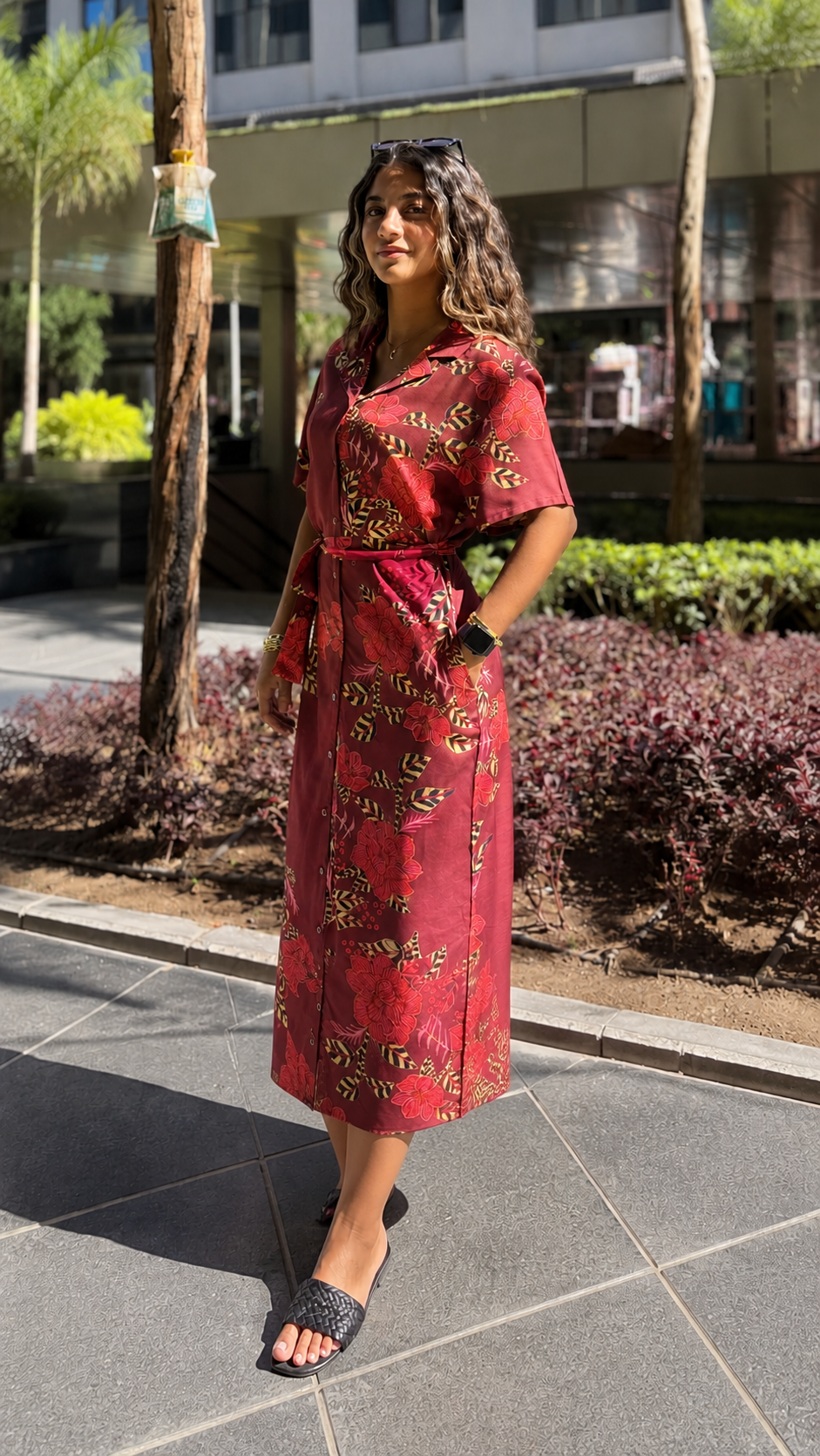 Image of Woman in a red patterned dress standing on a sidewalk with buildings and greenery in the background