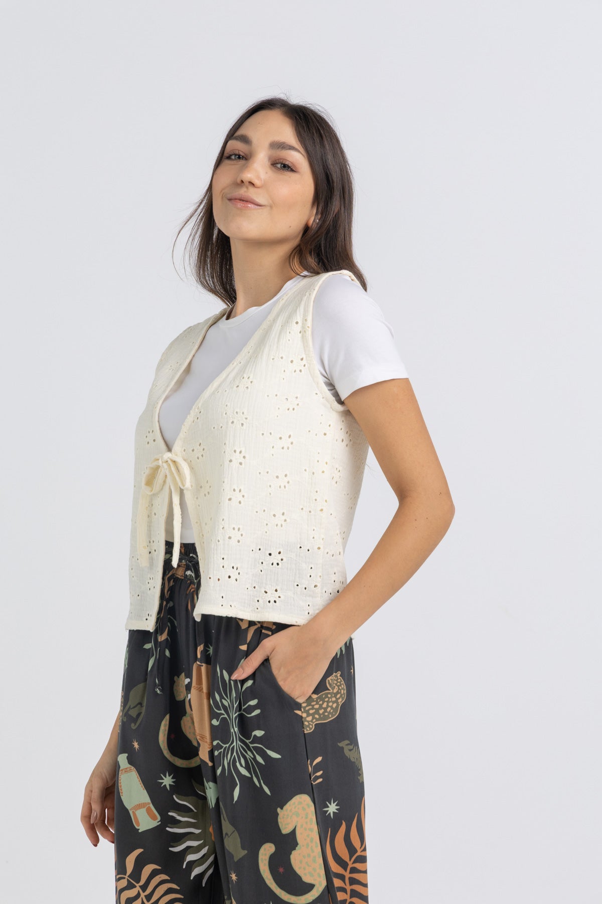 Woman wearing a white lace vest over a white shirt with black patterned pants on a light gray background